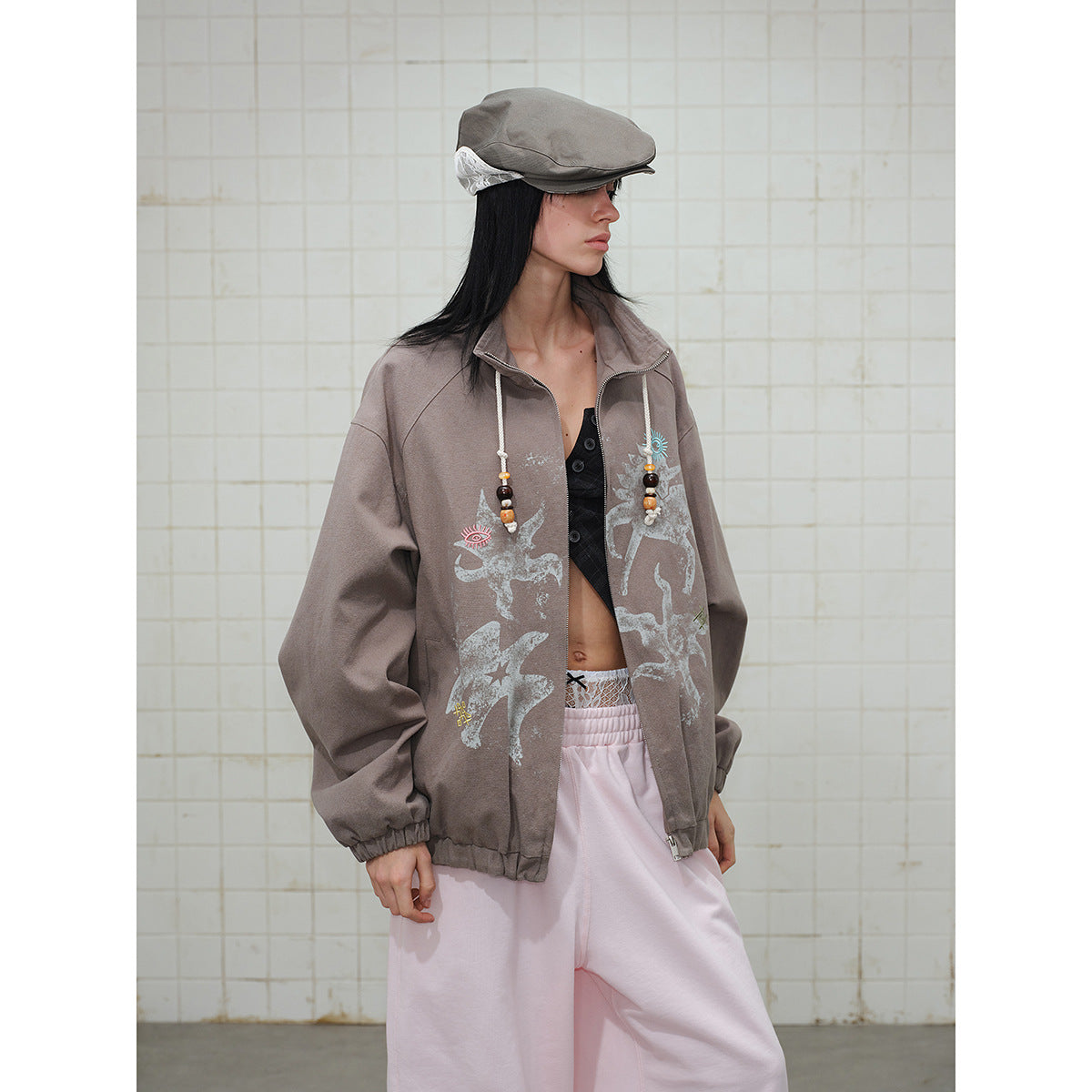 Model wearing a taupe zip-front jacket with pale graphic prints and beaded drawstrings, paired with pale pink pants.