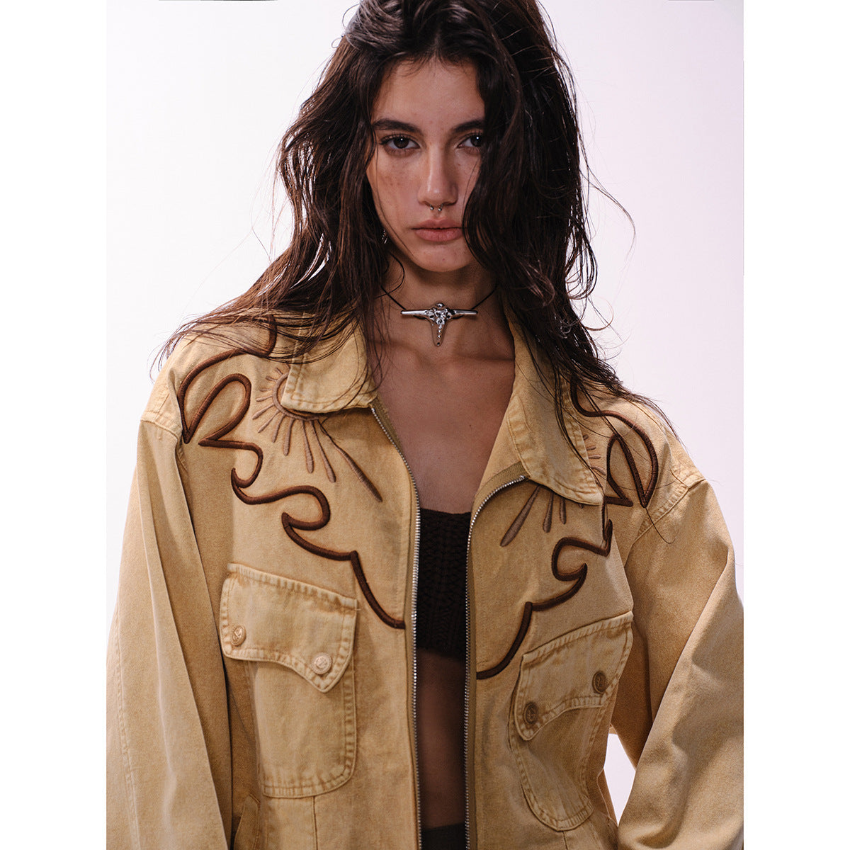 Close view of a tan jacket showing dark swirling embroidery, pointed collar, zipper and flap chest pockets.