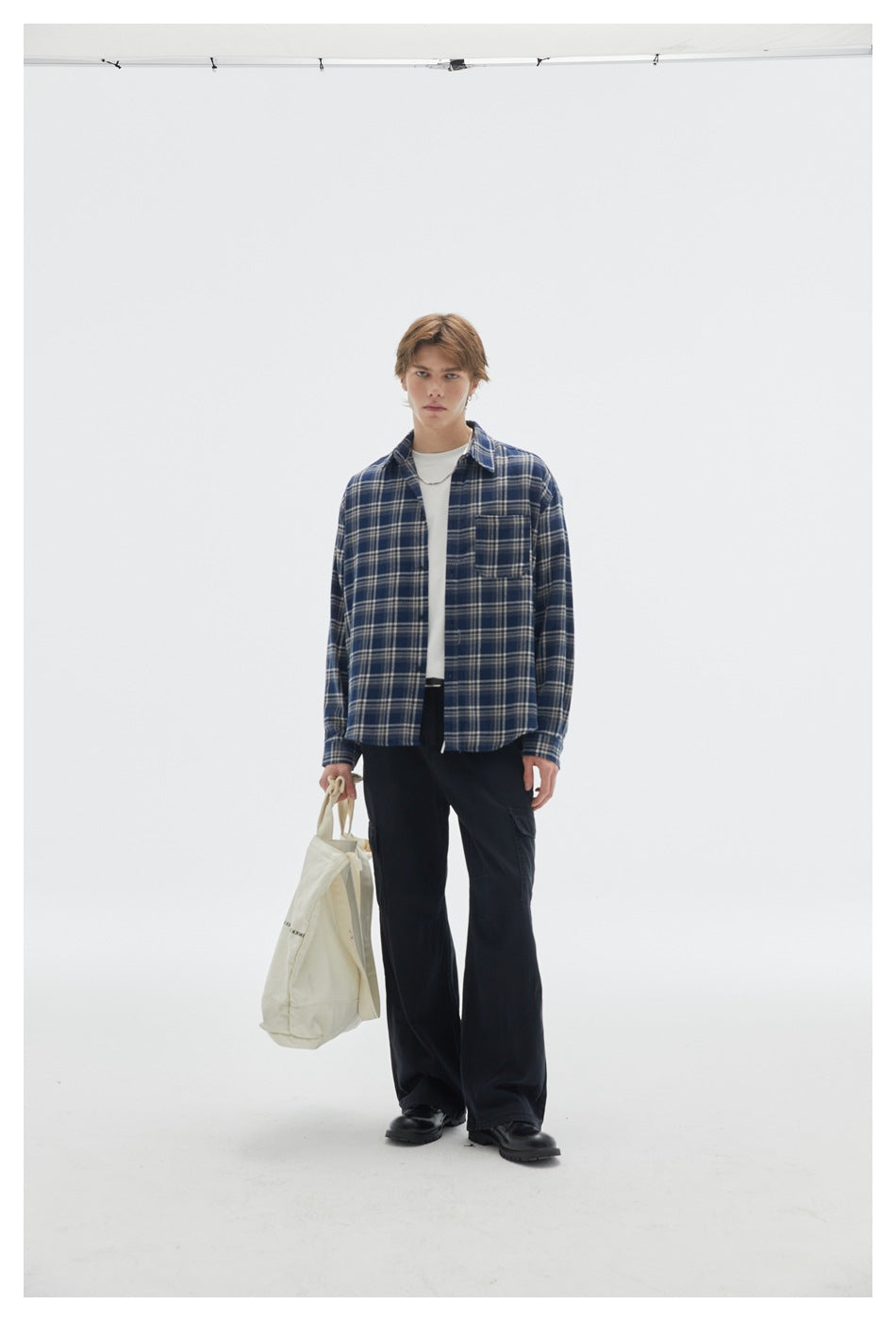 Model wearing black cargo pants with a blue plaid shirt and white tee, holding a cream tote bag