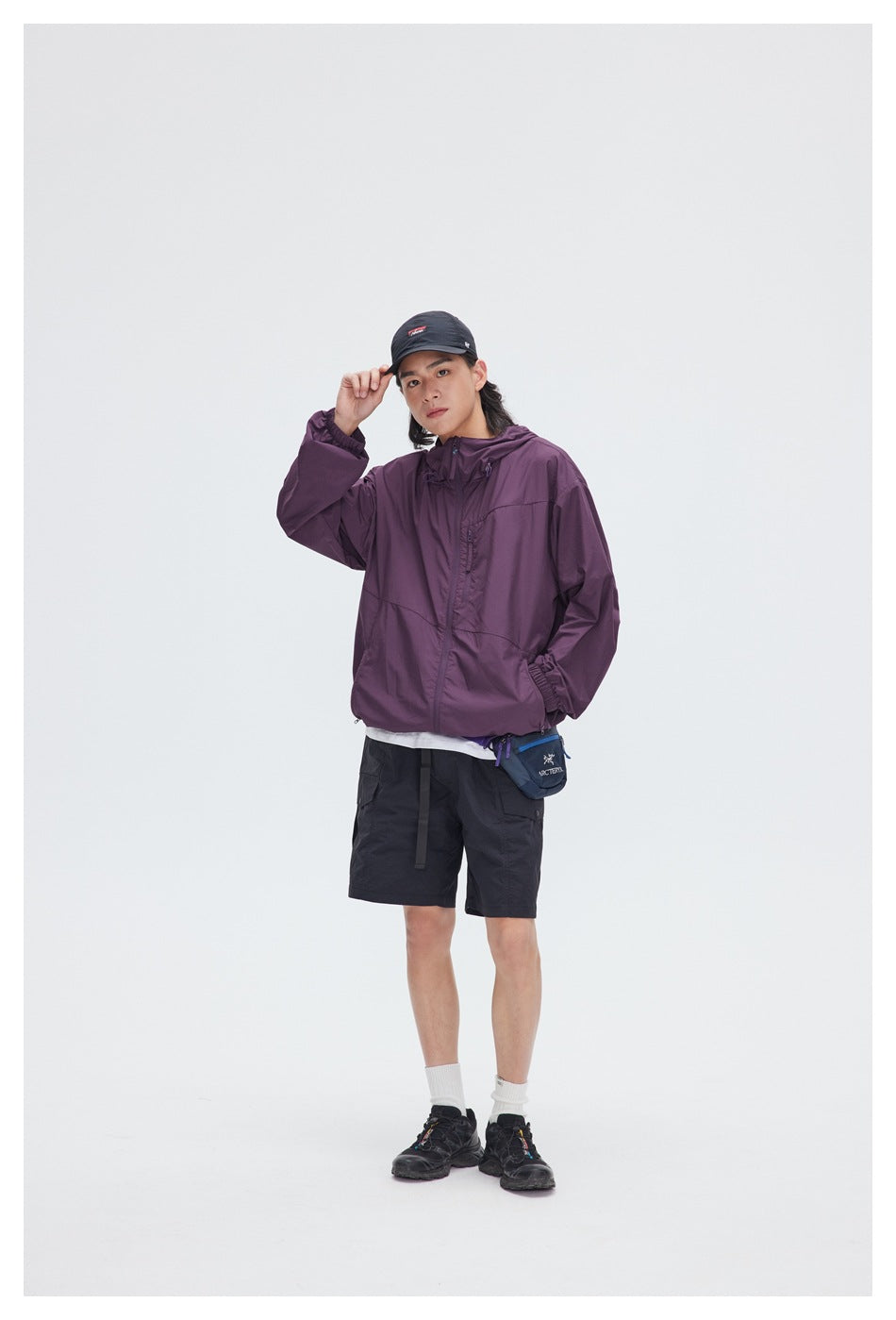 Model wearing a plum hooded zip jacket and black shorts, with a small blue crossbody bag.