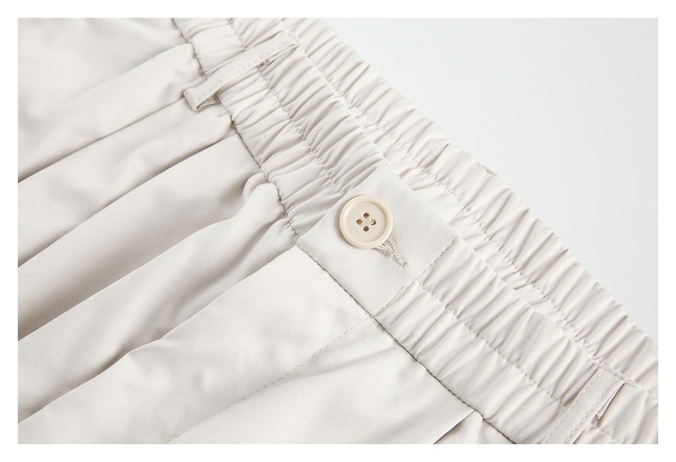 Close-up of off-white trousers showing the gathered elastic waistband, button, belt loop, and pleated front.