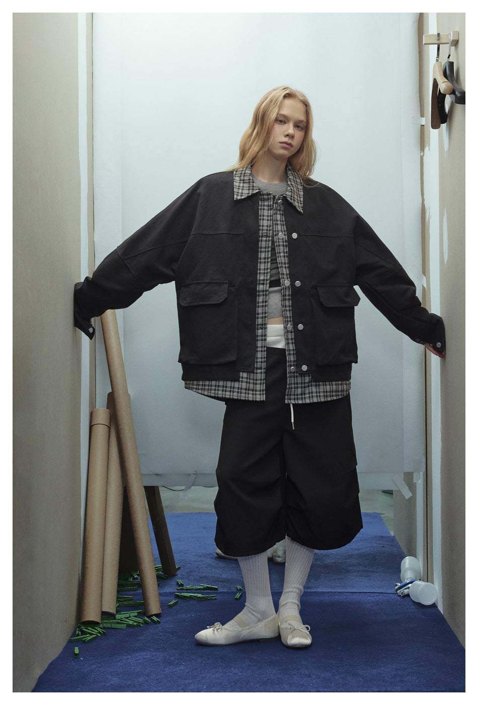 Model wearing a black oversized jacket with plaid collar and large flap pockets over a plaid layer, arms outstretched
