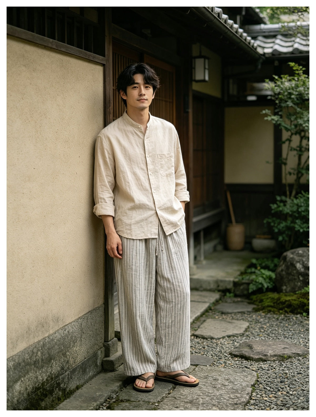 Man in striped wide-leg drawstring pants, beige shirt and sandals standing against a wall in a courtyard
