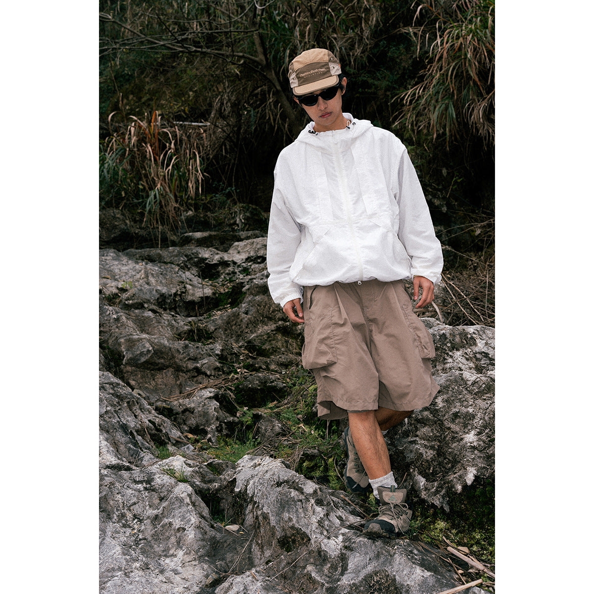 A man standing on rocky terrain wears a white hooded jacket and taupe cargo shorts.