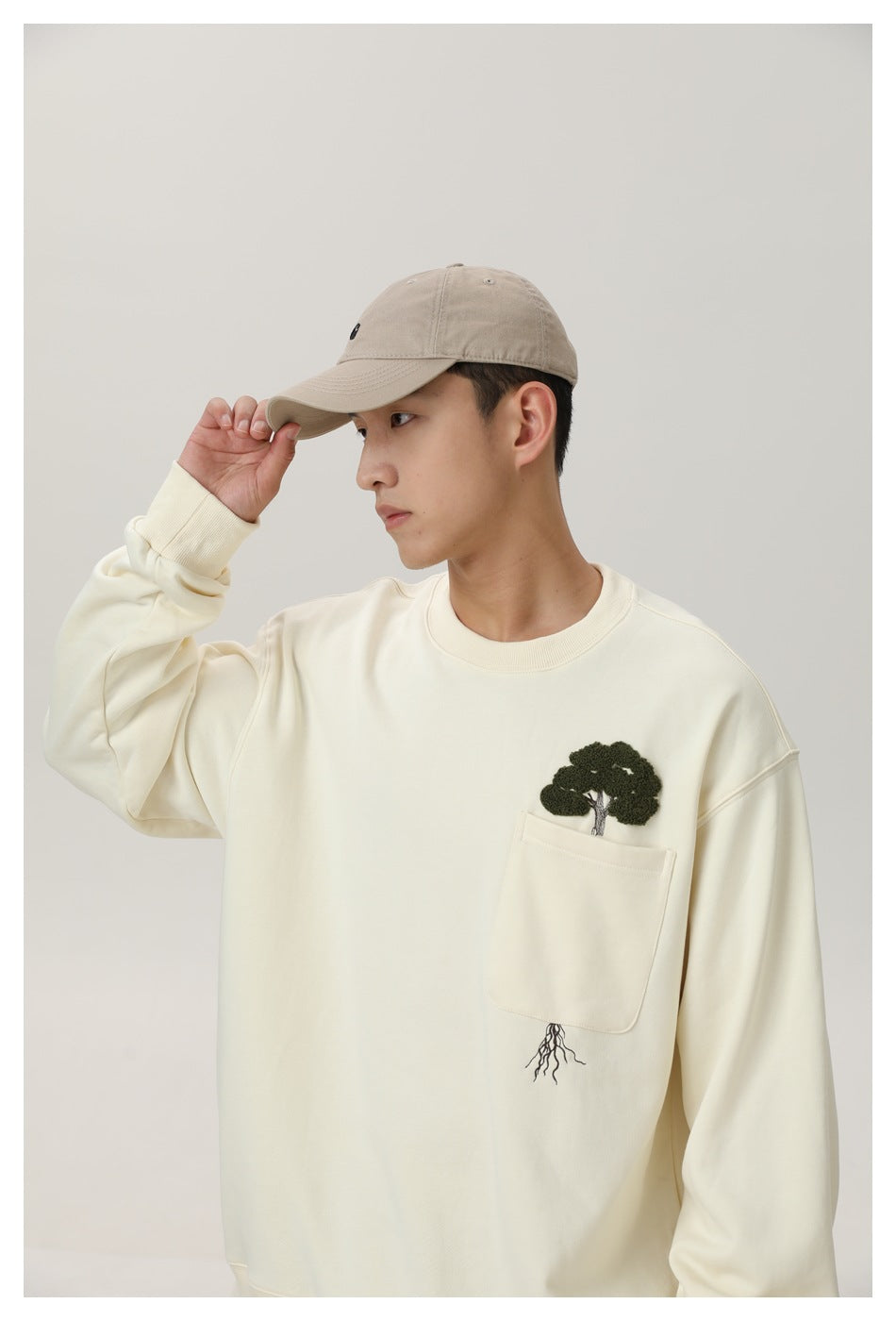 Side view of a model wearing an ivory sweatshirt with a pocket and embroidered tree motif.
