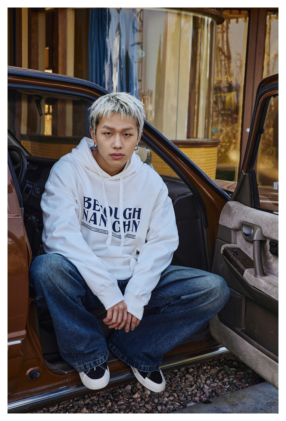 Model seated in an open car wearing a white graphic pullover hoodie and loose blue jeans.
