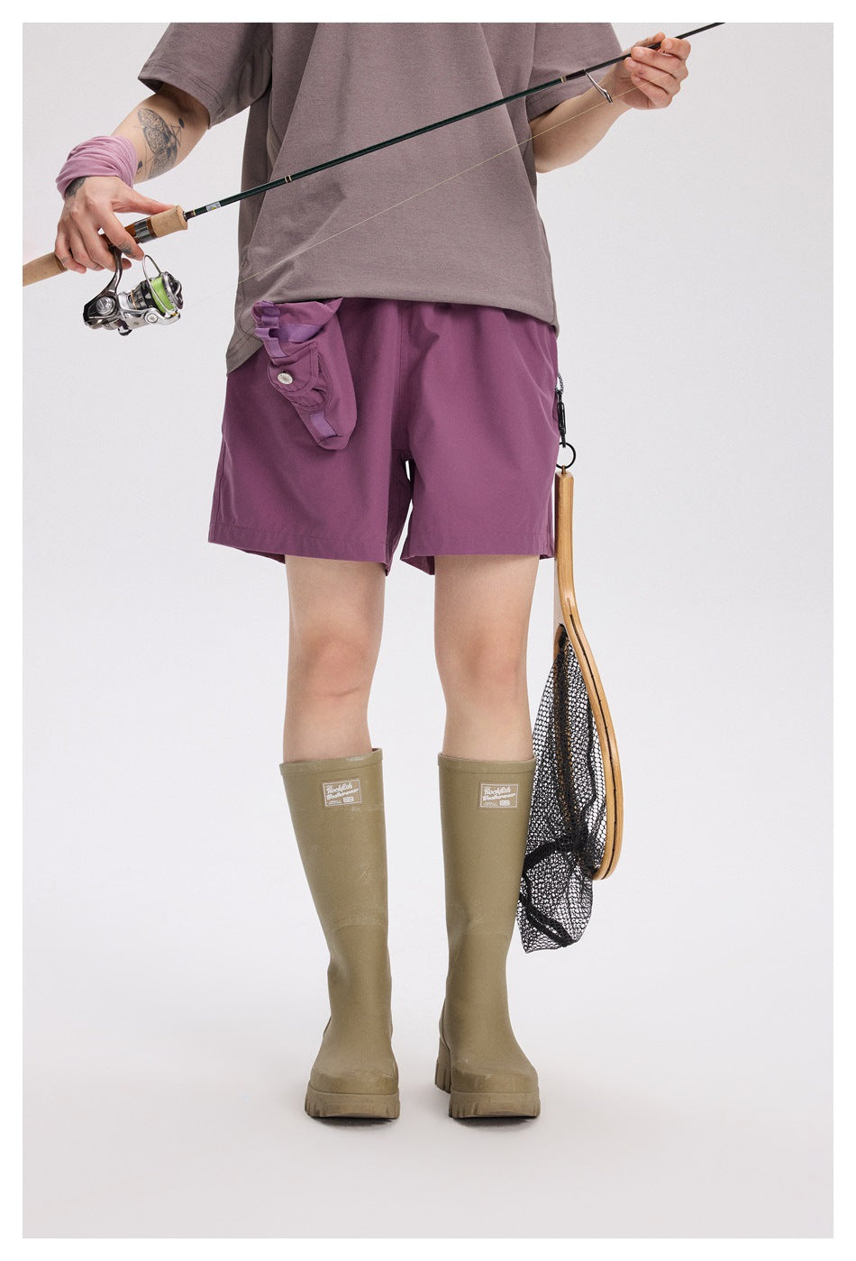Front view of a person wearing plum-purple shorts with a gray shirt and khaki boots, holding a fishing rod.