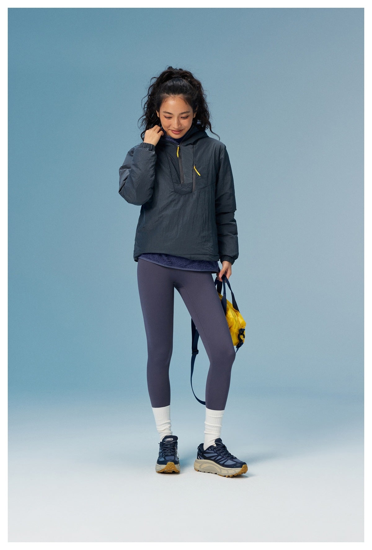 Woman modeling a charcoal hooded pullover with yellow zipper pulls and purple leggings, shown standing against a blue