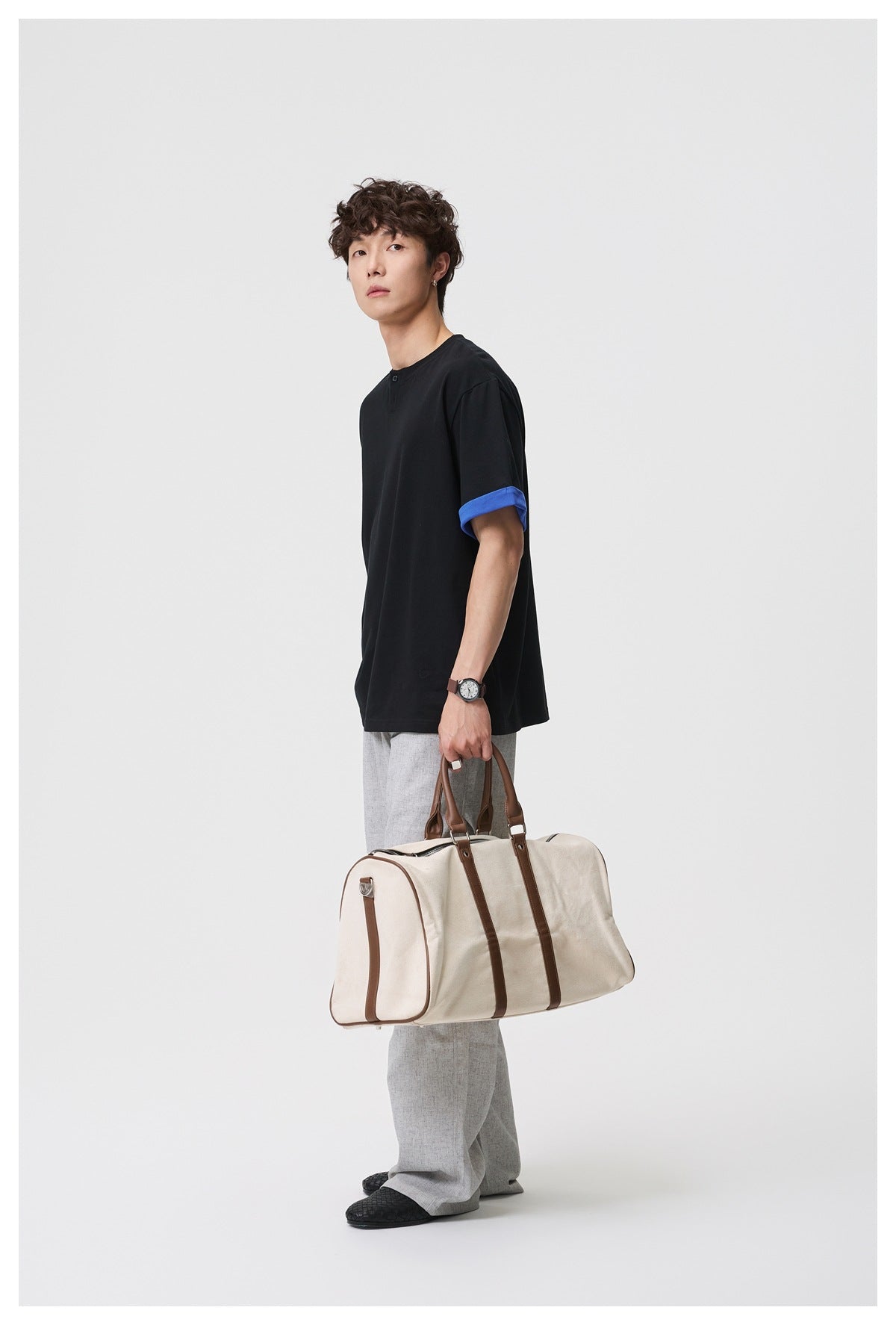 Model wearing a black short-sleeve henley T-shirt with a blue turned-up cuff, light gray trousers, and carrying a cream bag.
