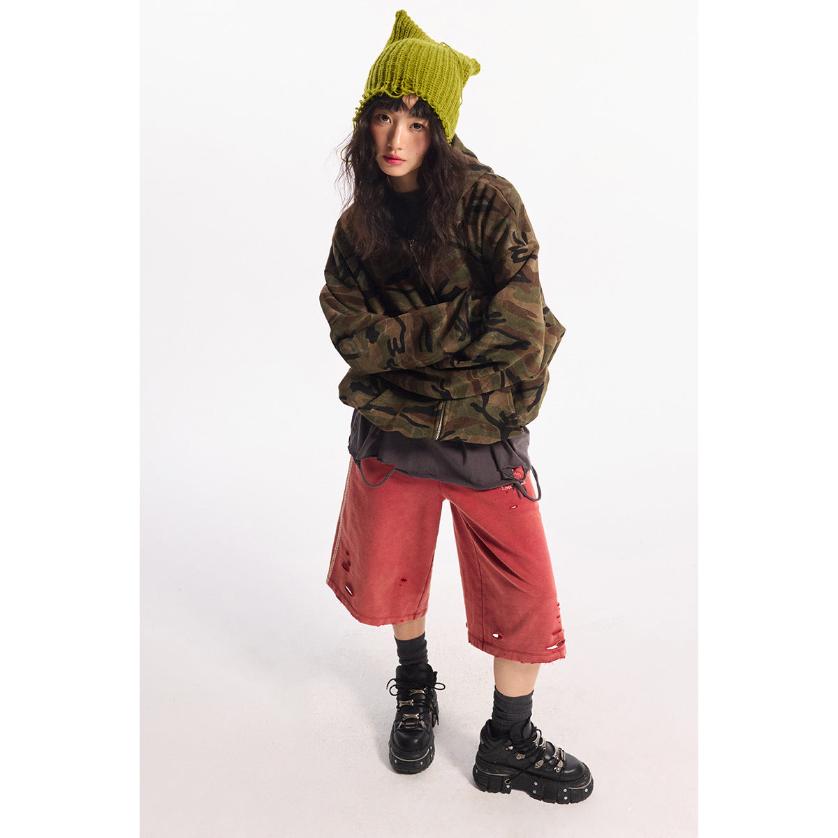 Model wearing an oversized brown and olive camouflage zip jacket, lime knit beanie and distressed red wide-leg shorts.