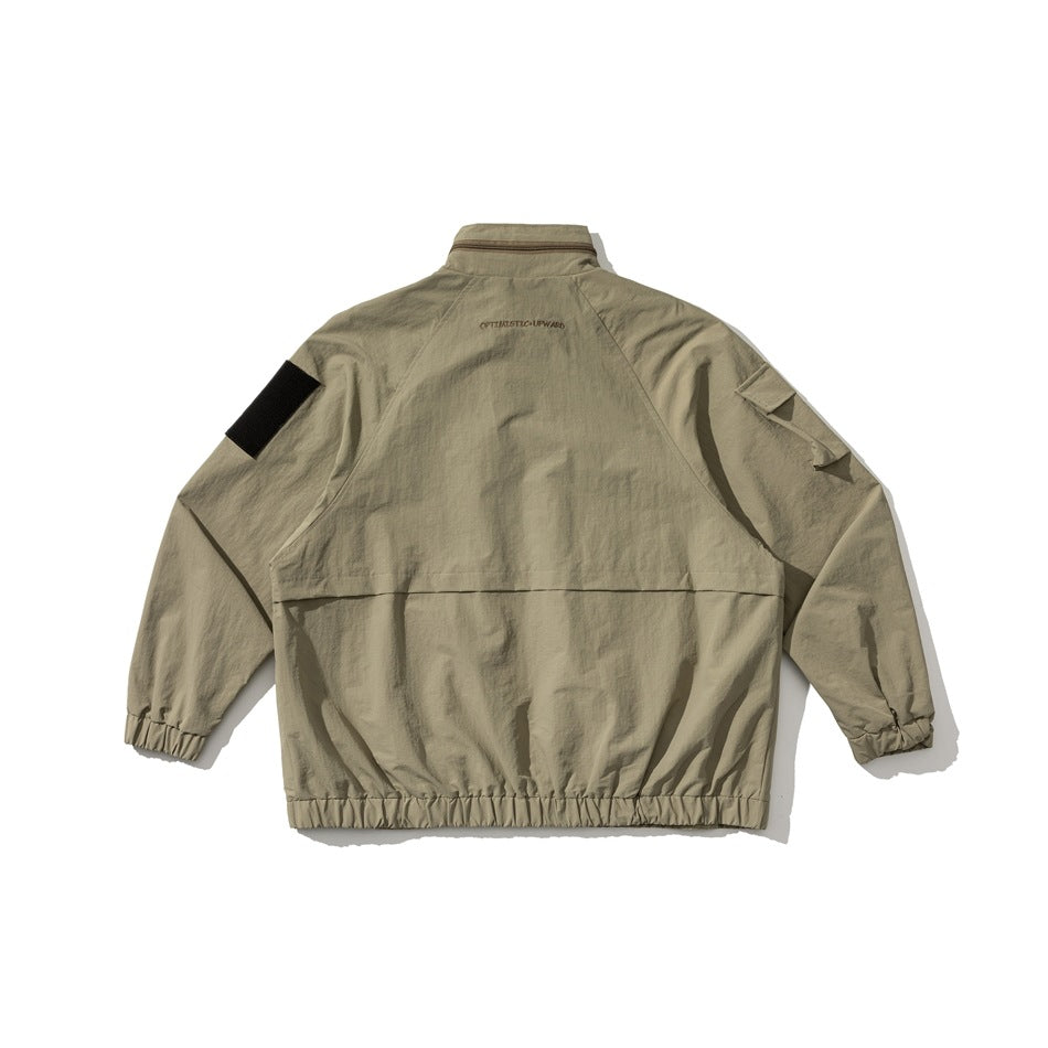 Back view of a khaki utility jacket with a high collar, raglan sleeves, black upper-sleeve patches, and an elastic hem.