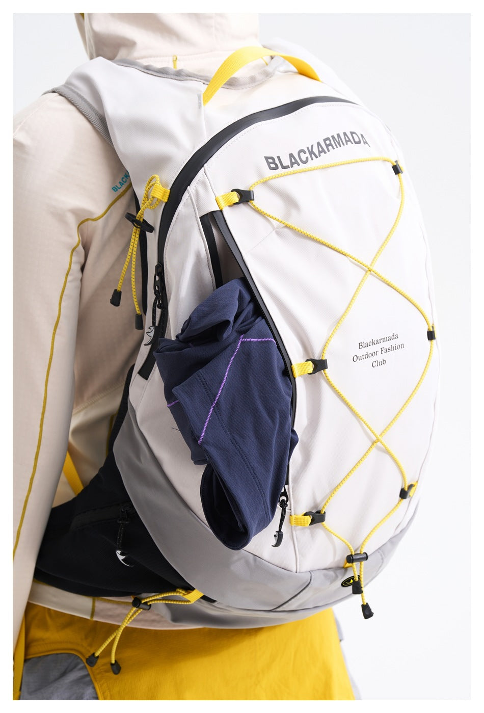 Close-up of a white hiking backpack with yellow cord lacing and a dark garment in its side pocket, worn over a pale jacket.
