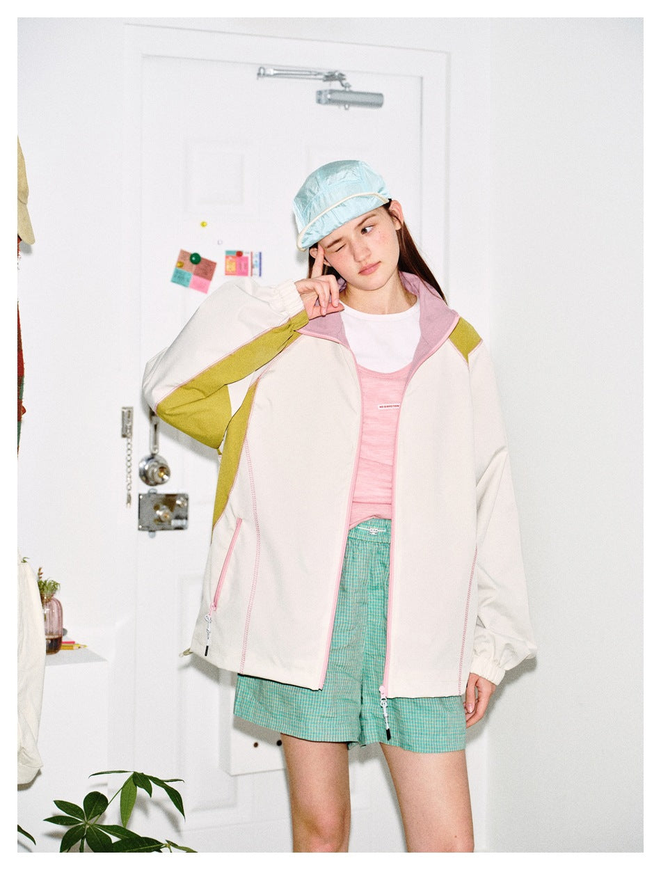 Model wearing the beige color-block jacket open over a pink top and green checked shorts, with a light blue cap