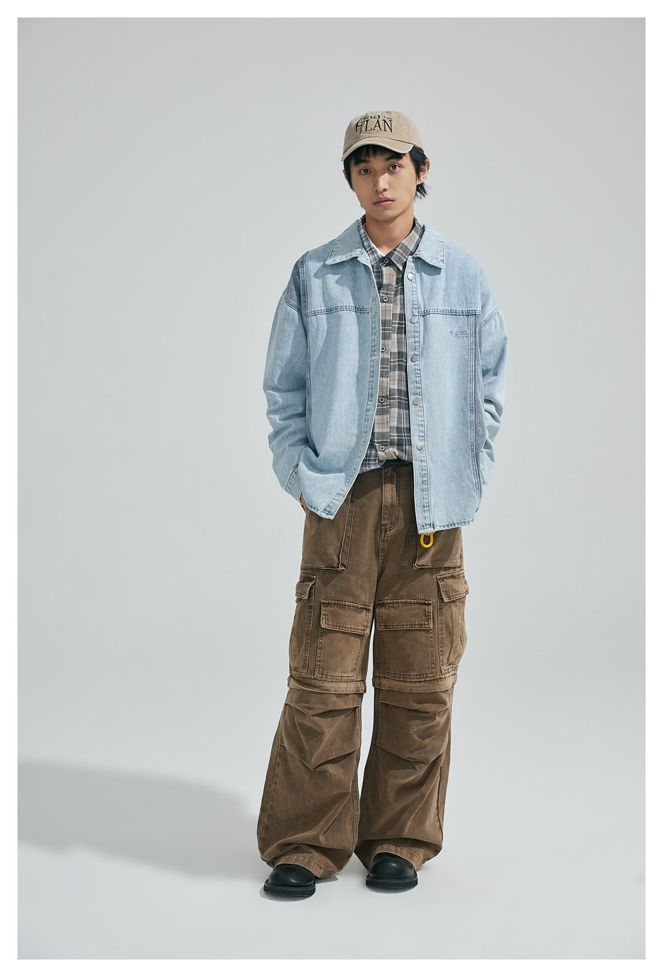 Model wearing a light blue denim jacket open over a plaid shirt with brown cargo pants and a cap
