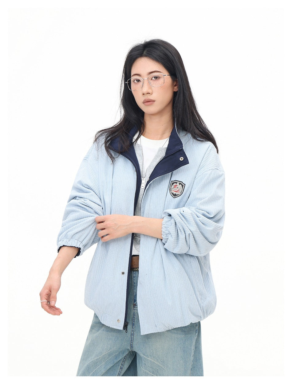 Model wearing a pale blue ribbed zip jacket with a navy collar and chest crest, layered over a gray top and jeans.
