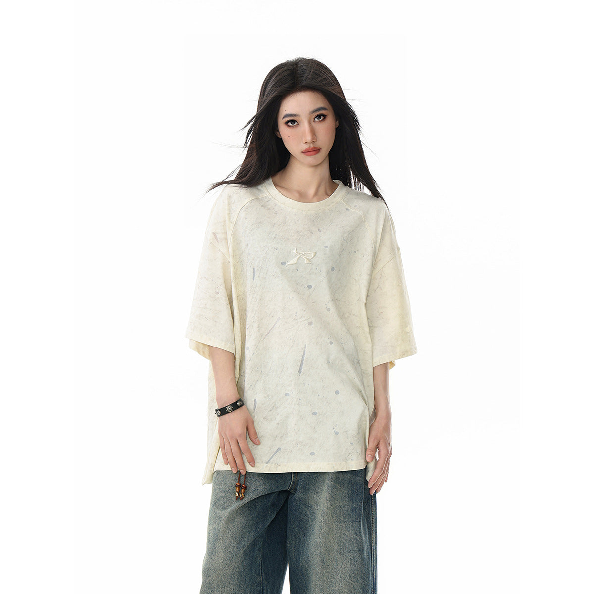 Model wearing an oversized ivory, gray-speckled T-shirt with a small chest emblem and loose dark blue jeans, front view.