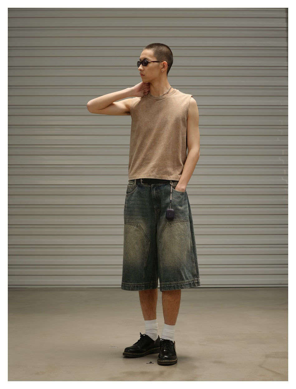 Three-quarter view of a man wearing a tan sleeveless top and loose, faded dark denim shorts.
