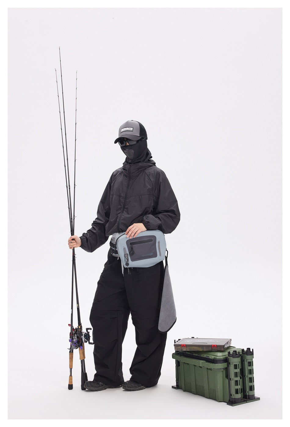 A masked person wears a black hooded jacket and trousers with a gray fishing bag, holding fishing rods beside tackle cases.