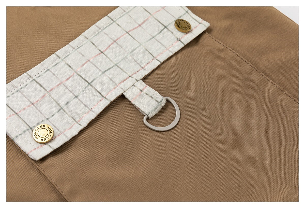 Close-up of a tan jacket pocket with a cream plaid flap, brass buttons and a metal D-ring.