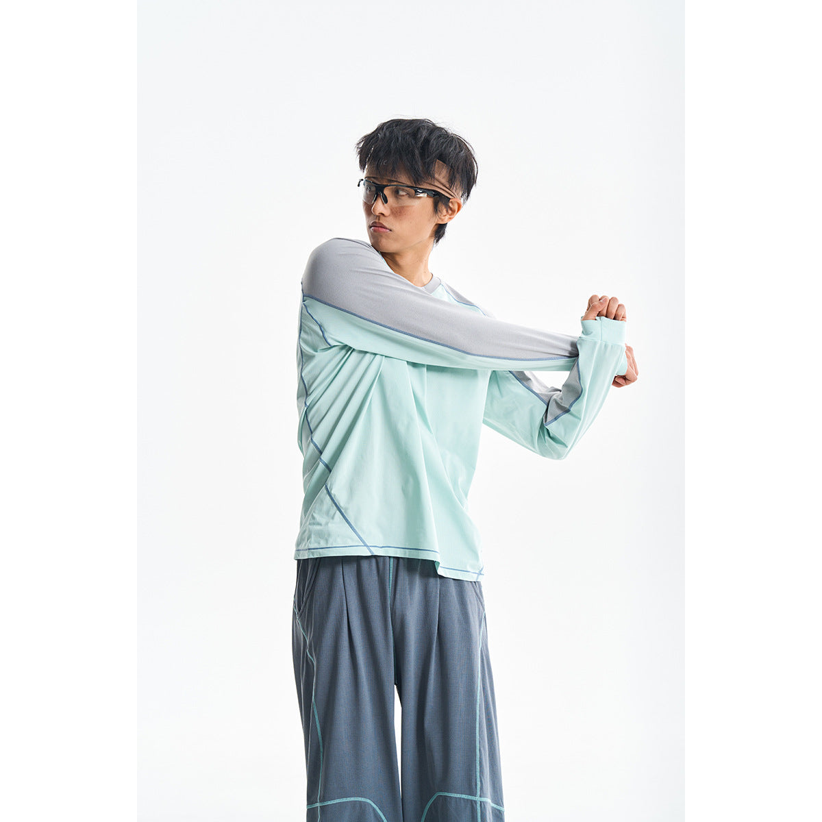 Model stretching in a pale aqua long-sleeve top with gray shoulder panels and loose dark gray pants.