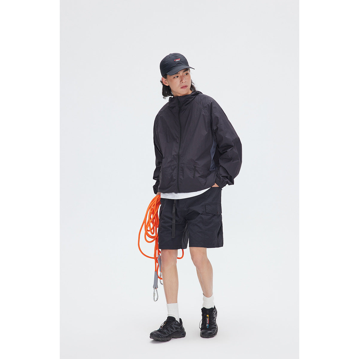 Full-length model wearing a black hooded zip jacket, black shorts, cap, and hiking shoes.