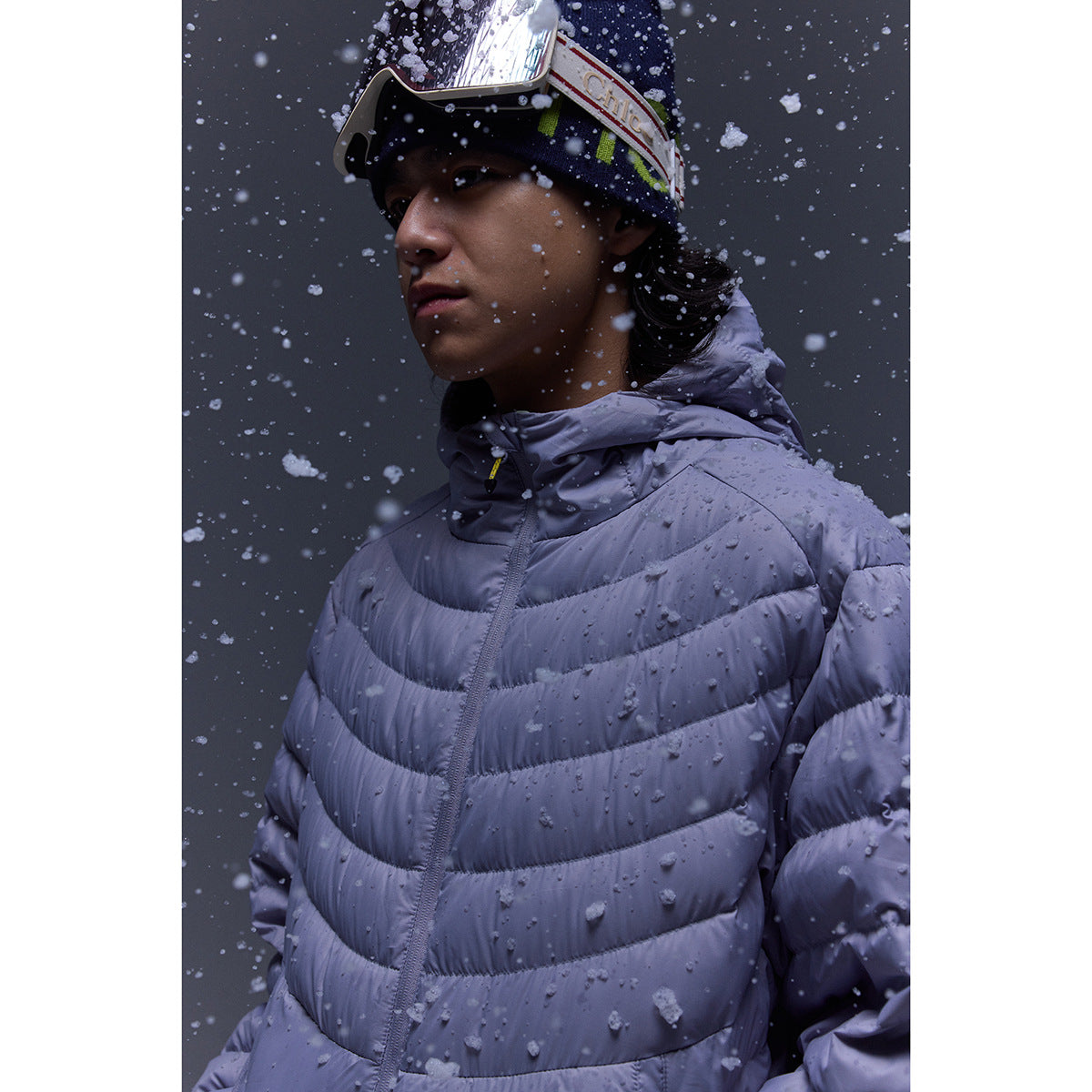 Close-up of a model in a lavender hooded puffer jacket as snow falls around them.