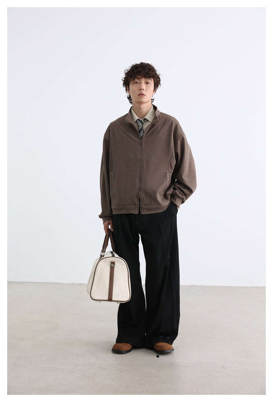 Model wearing a brown zip-front jacket with a stand collar, black trousers, and carrying a cream bag.