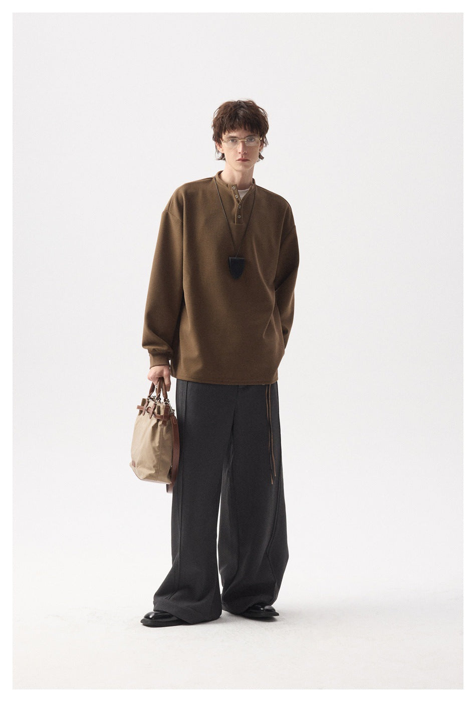 Model wearing brown Henley pullover with wide dark gray trousers, holding a beige tote bag