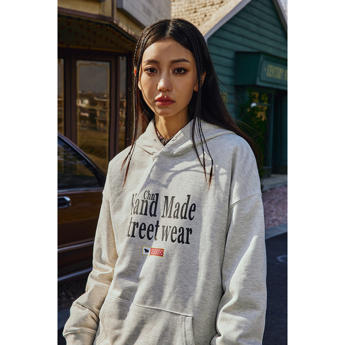 Model wearing a light gray pullover hoodie with a large dark front graphic, kangaroo pocket, and drawstring hood.