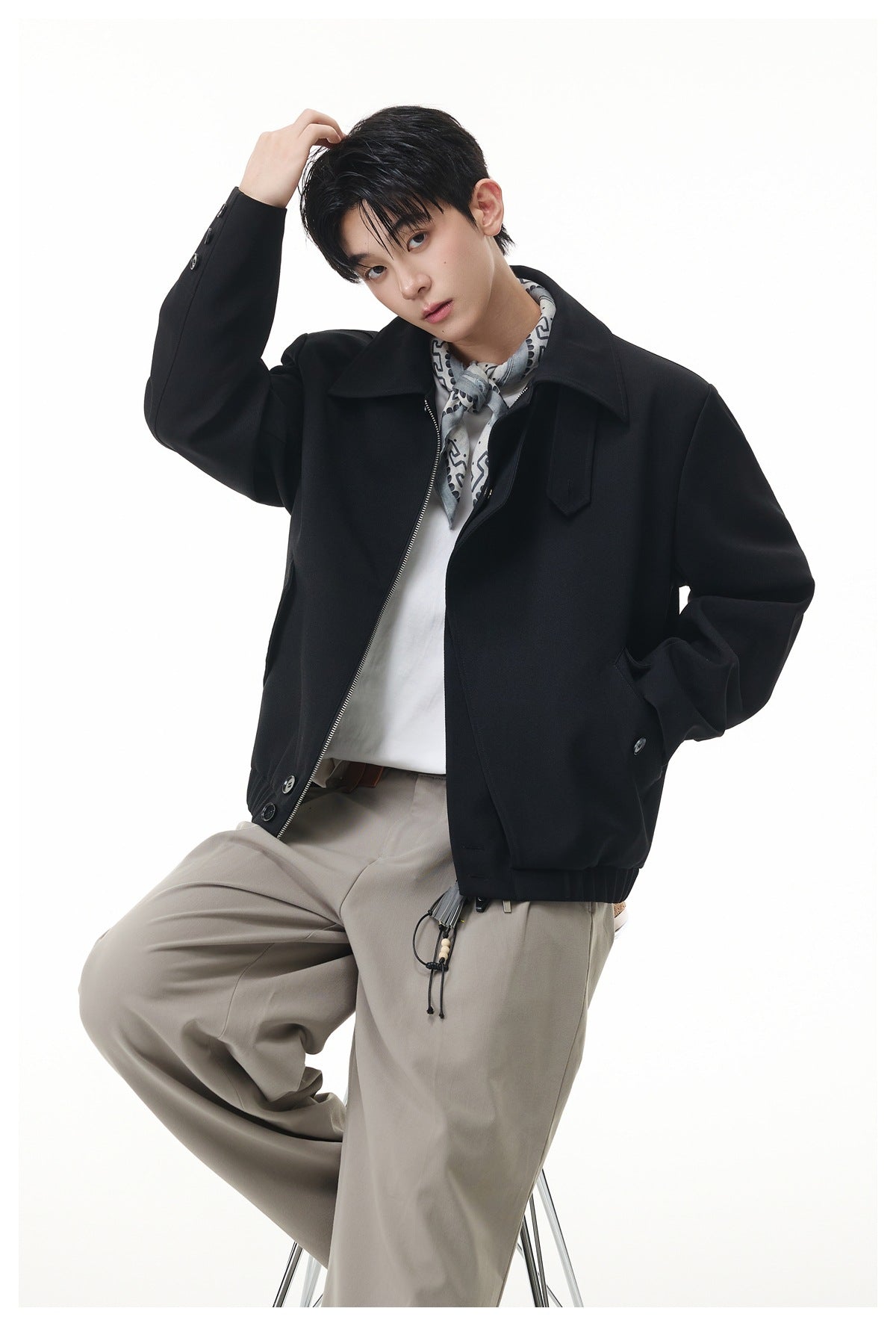 Seated model wearing an open black jacket over a white shirt with beige trousers and a patterned neck scarf.