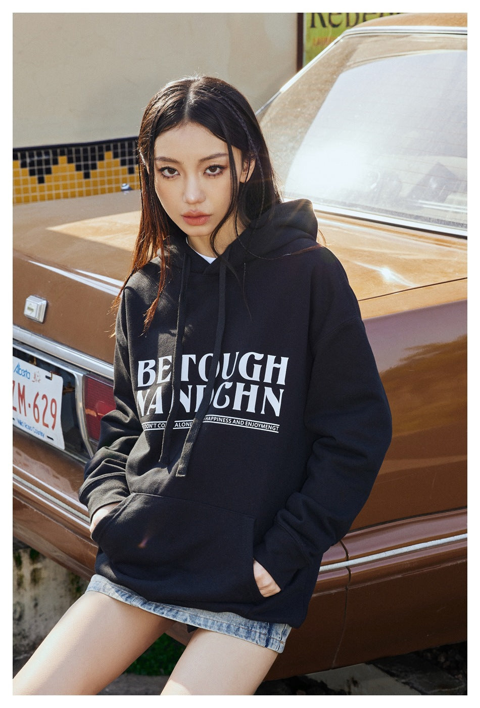 Model wears a black oversized graphic hoodie with a kangaroo pocket and denim shorts, posed beside a car.