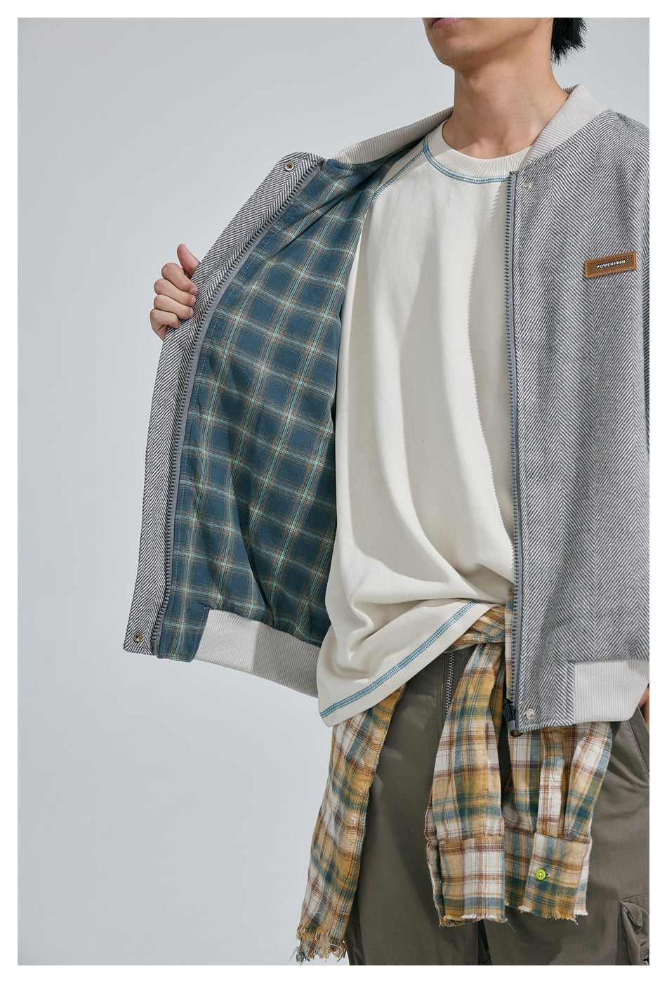 Close view of a model holding open a gray herringbone bomber jacket to show its blue plaid lining.