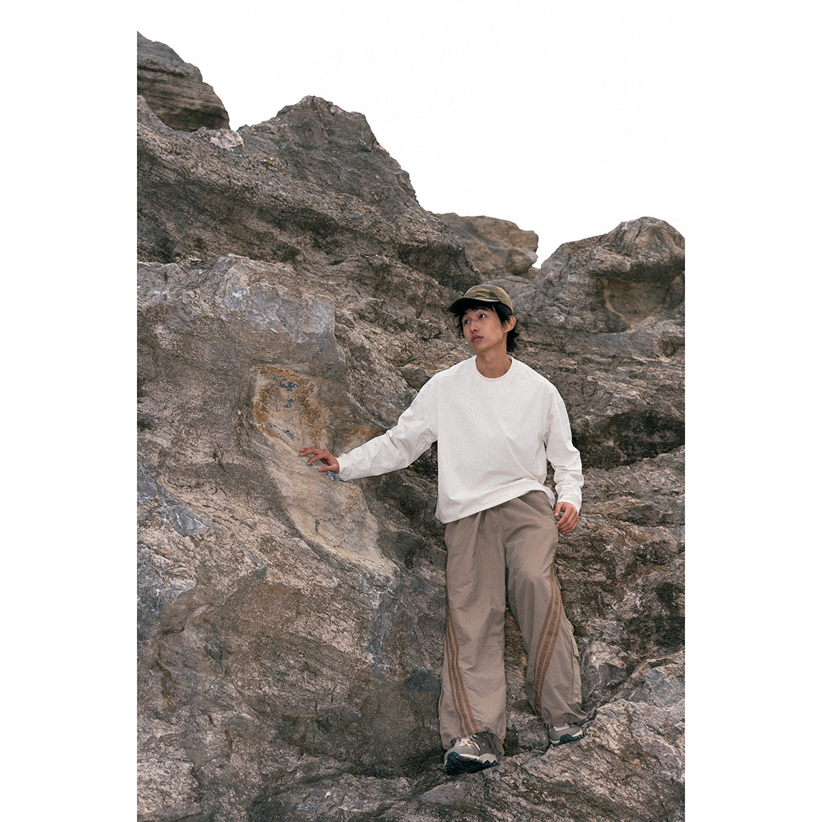 Model wearing an off-white long-sleeve shirt and taupe wide-leg trousers among rocky cliffs.