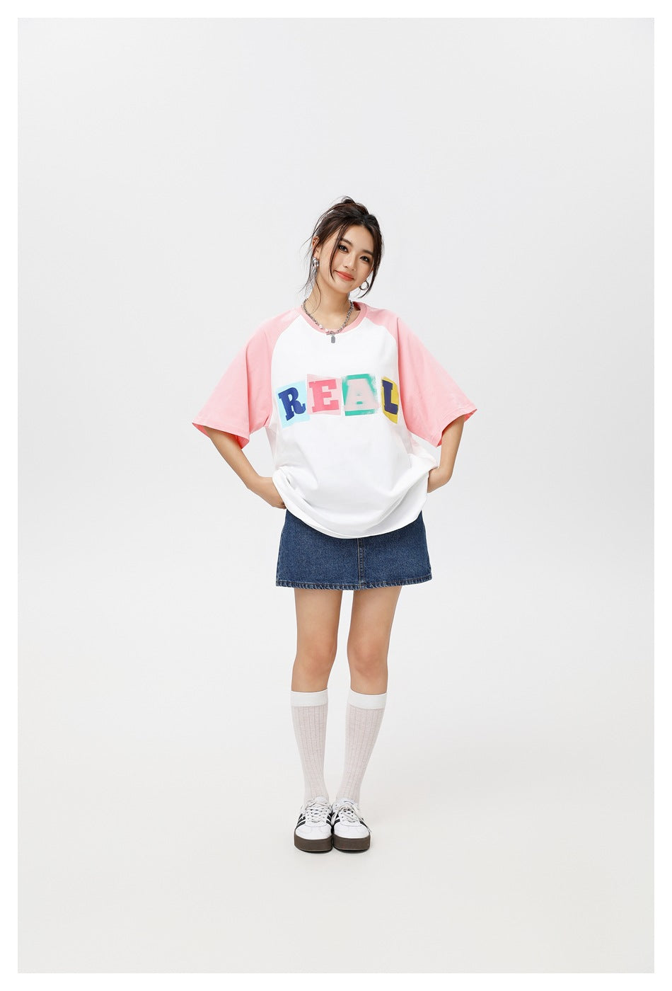 Full-length model wearing a white T-shirt with pink raglan sleeves and colorful REAL lettering, with a denim skirt.
