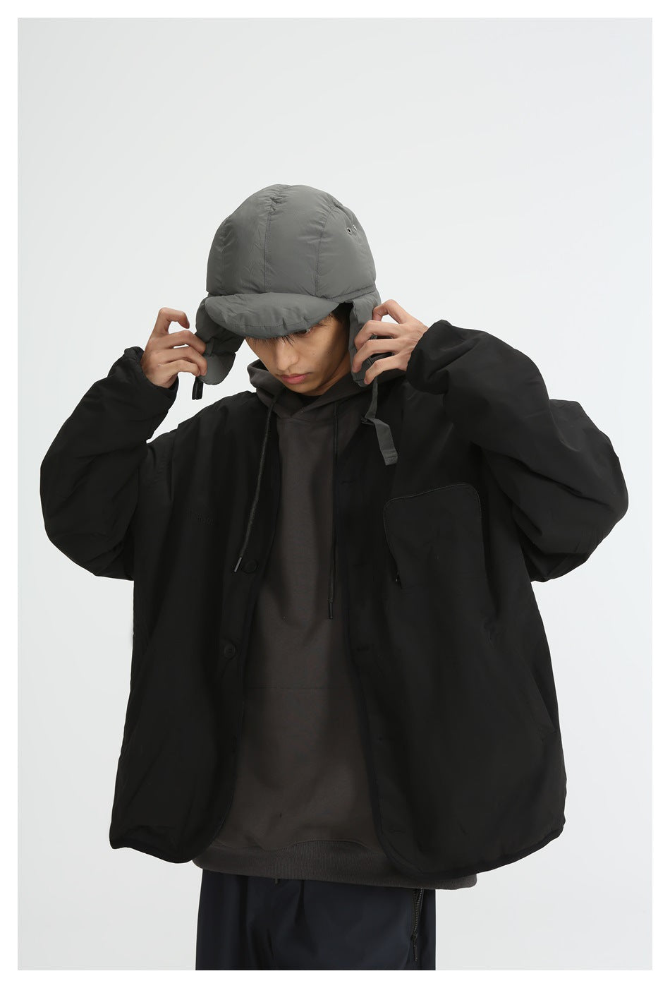 Front view of a model wearing a black oversized jacket over a dark hoodie, adjusting a gray padded earflap cap.