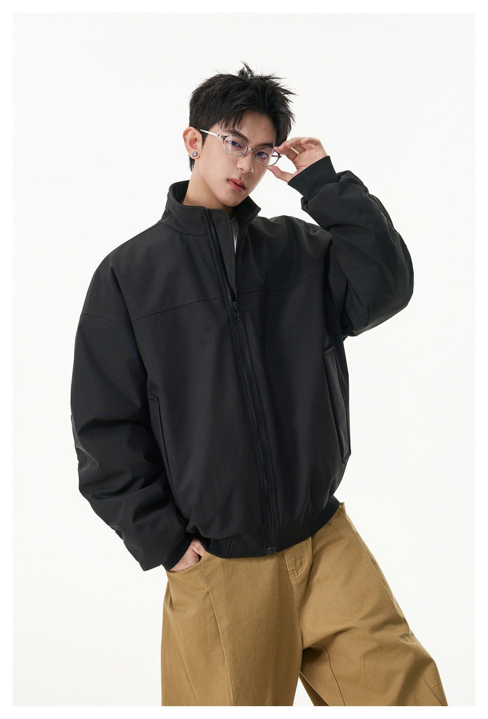 Model wearing a black oversized zip-up jacket with a stand collar and front pockets, shown with tan trousers.