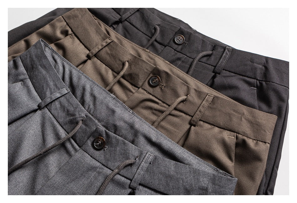 Three pairs of pleated trousers in charcoal gray, brown, and black arranged diagonally, showing their waistbands and buttons.