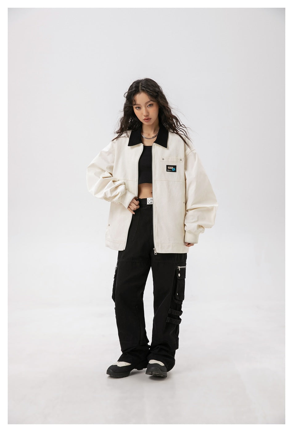 Model wearing an open beige work jacket with black collar over a black top and black cargo pants, full-length view