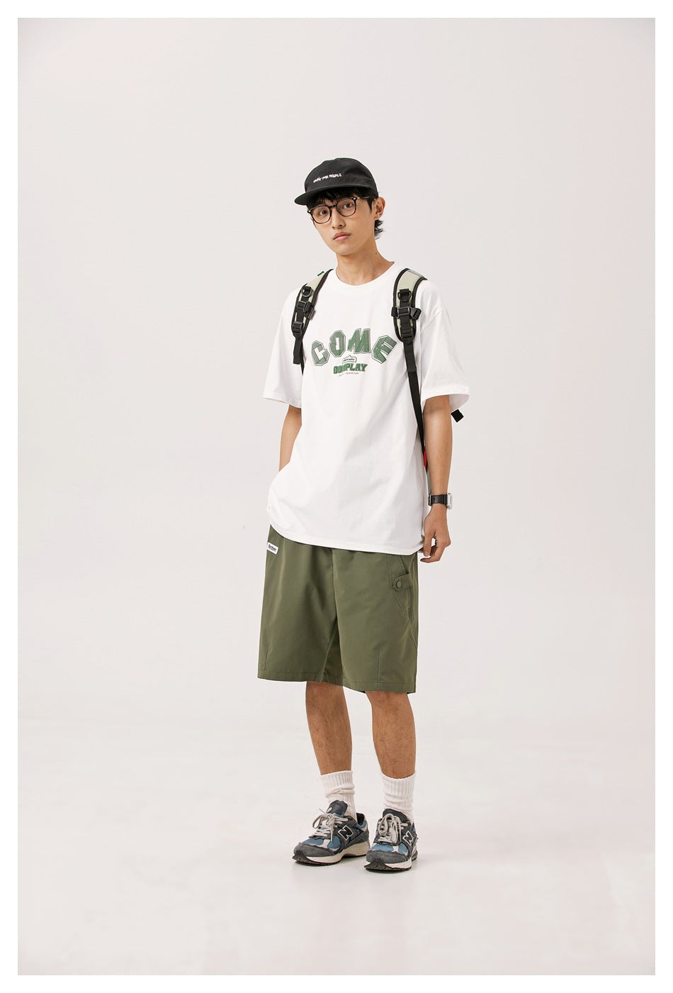 Full-length view of model in white graphic T-shirt, olive shorts, backpack, cap and sneakers