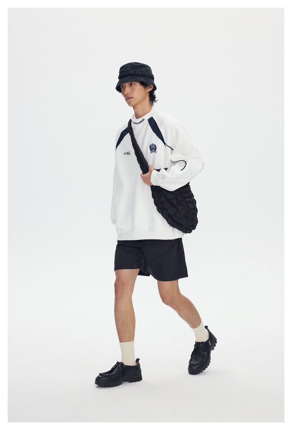 Person walking in a white crewneck sweatshirt with navy shoulder panels, black shorts, bucket hat and black quilted shoulder