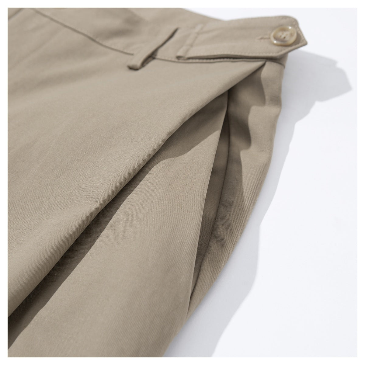 Close-up of taupe trousers showing a buttoned waistband tab, belt loop and folded fabric.