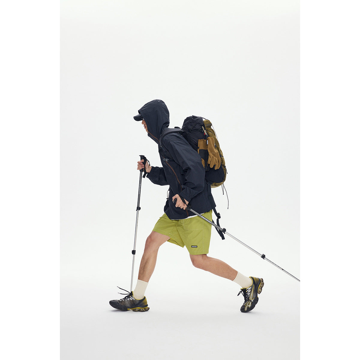 Model wearing a dark hooded jacket and lime-green shorts with a hiking backpack and trekking poles.