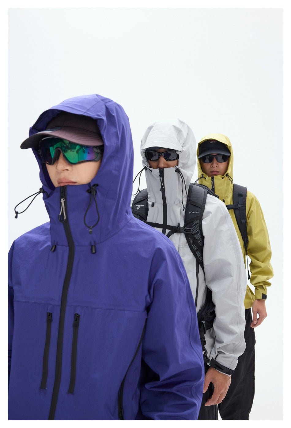 Close-up of three hikers wearing purple, light gray, and yellow hooded jackets with sunglasses and backpacks.