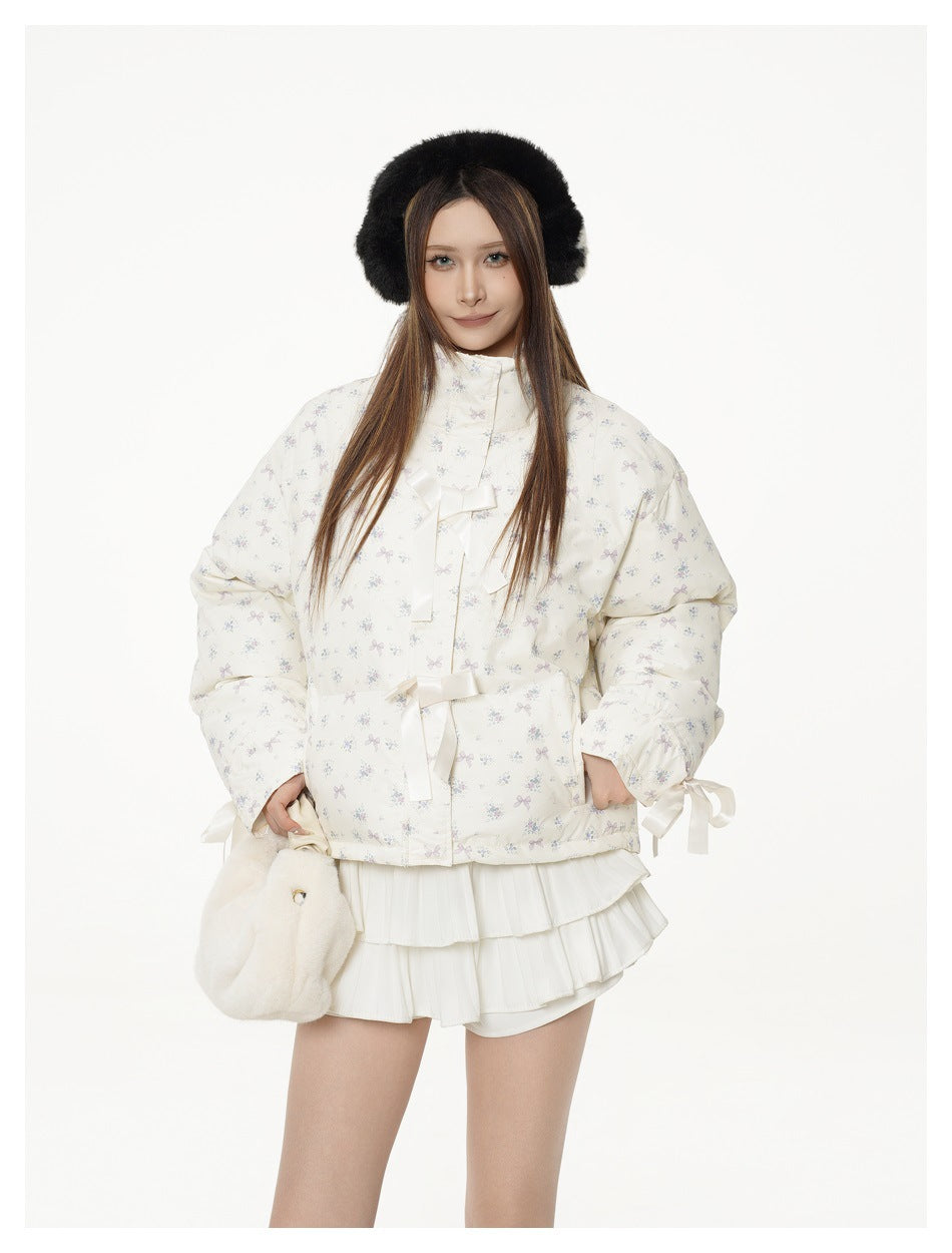 Model wearing a cream floral-print quilted jacket with ribbon bows, a white pleated mini skirt and a black headband
