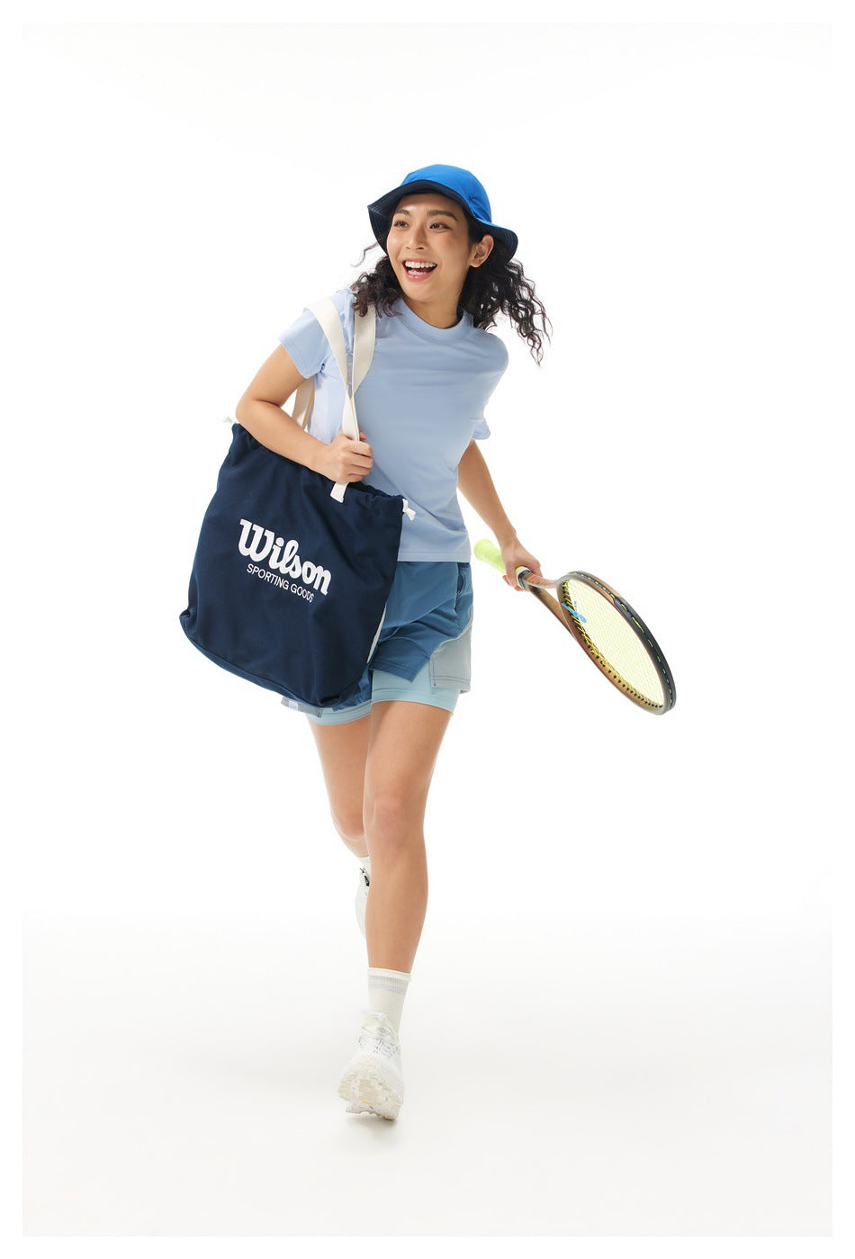 Model in a light blue T-shirt and blue layered shorts carries a navy Wilson tote and a tennis racket.