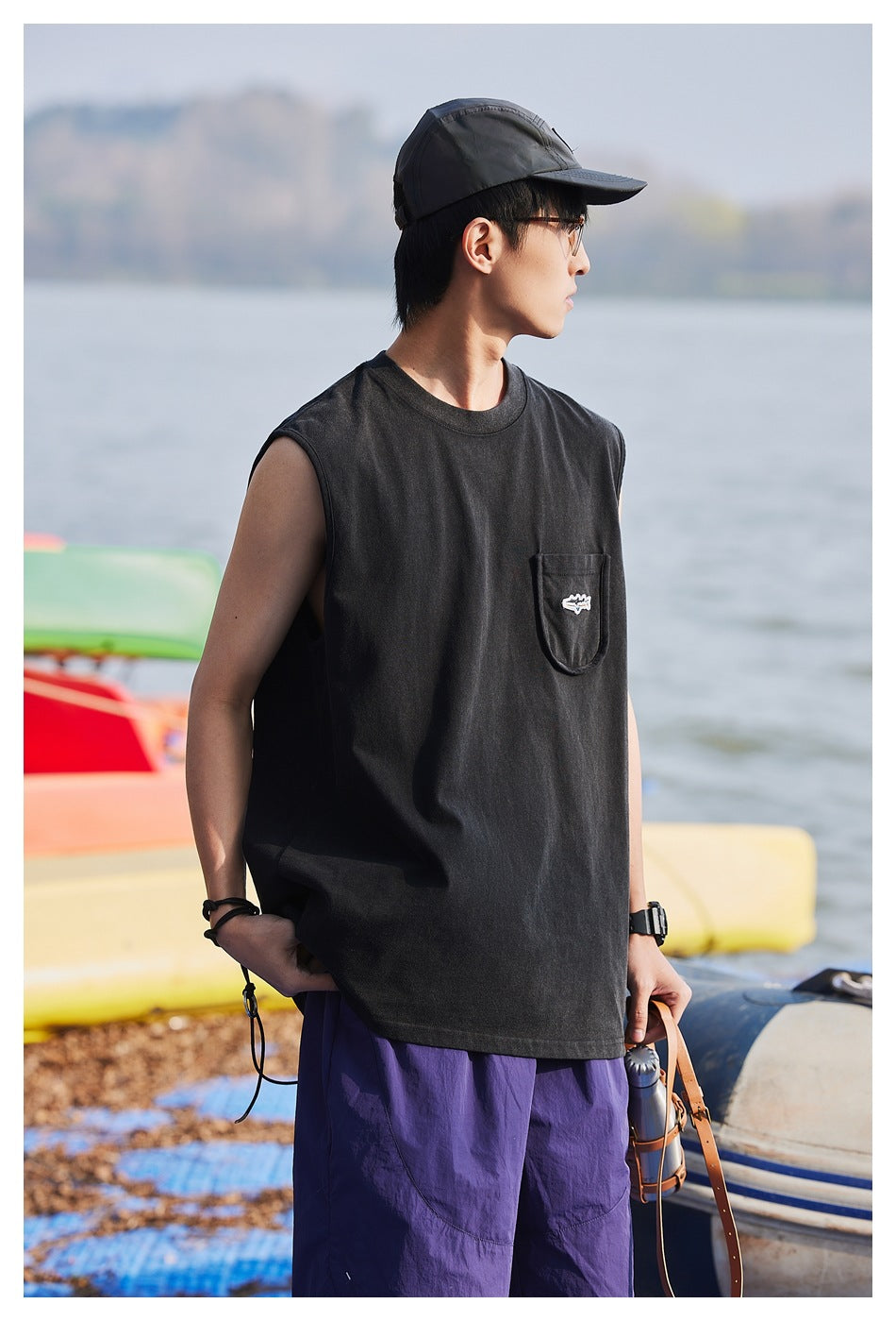 Side view of a model wearing a black sleeveless pocket shirt and purple shorts by the water.