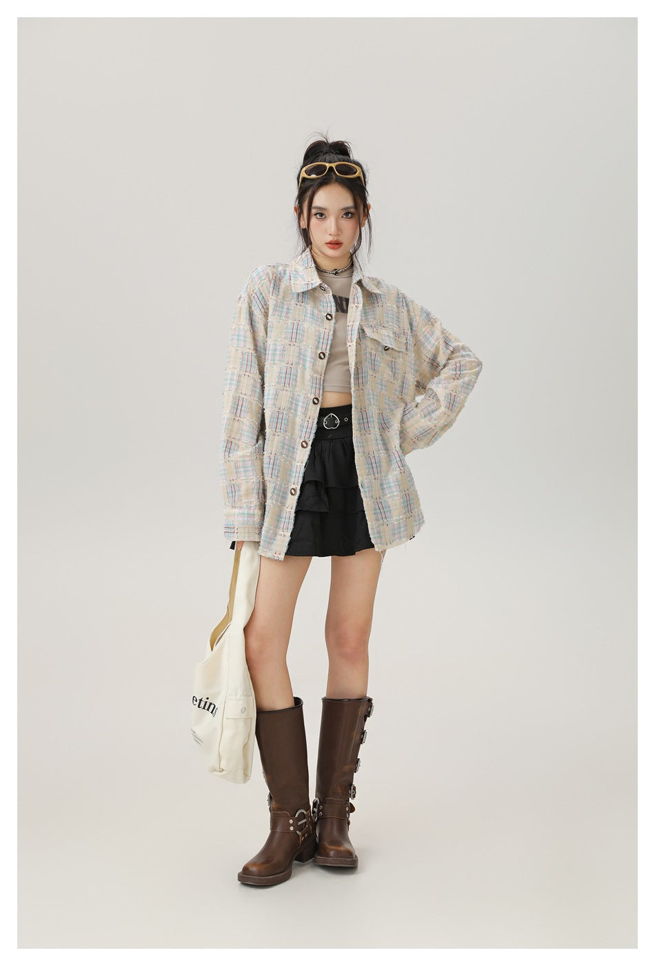 Model wearing open beige plaid fringed shirt over a cropped top, black ruffle skirt and brown boots, front view