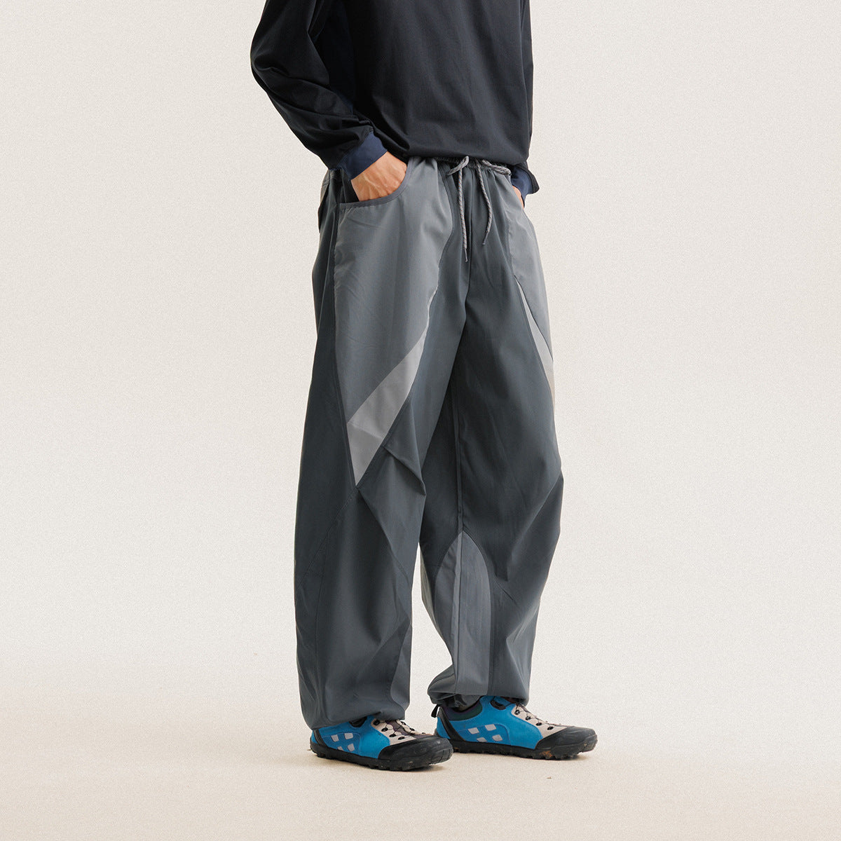 Model wearing loose charcoal-gray pants with contrasting curved panels and a drawstring waist, shown from torso to shoes.