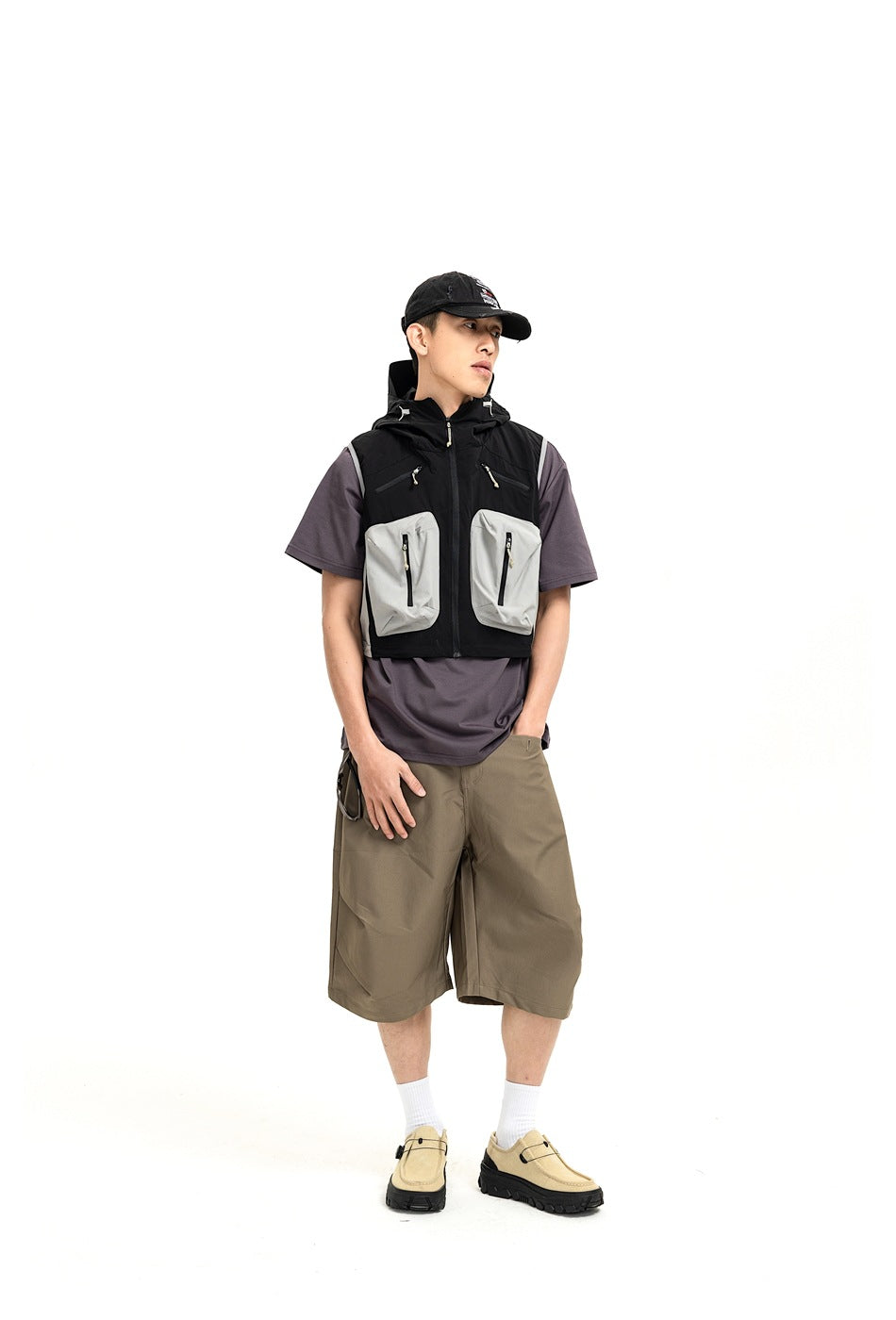Model wearing a black hooded utility vest with light gray pockets over a charcoal T-shirt and olive shorts.