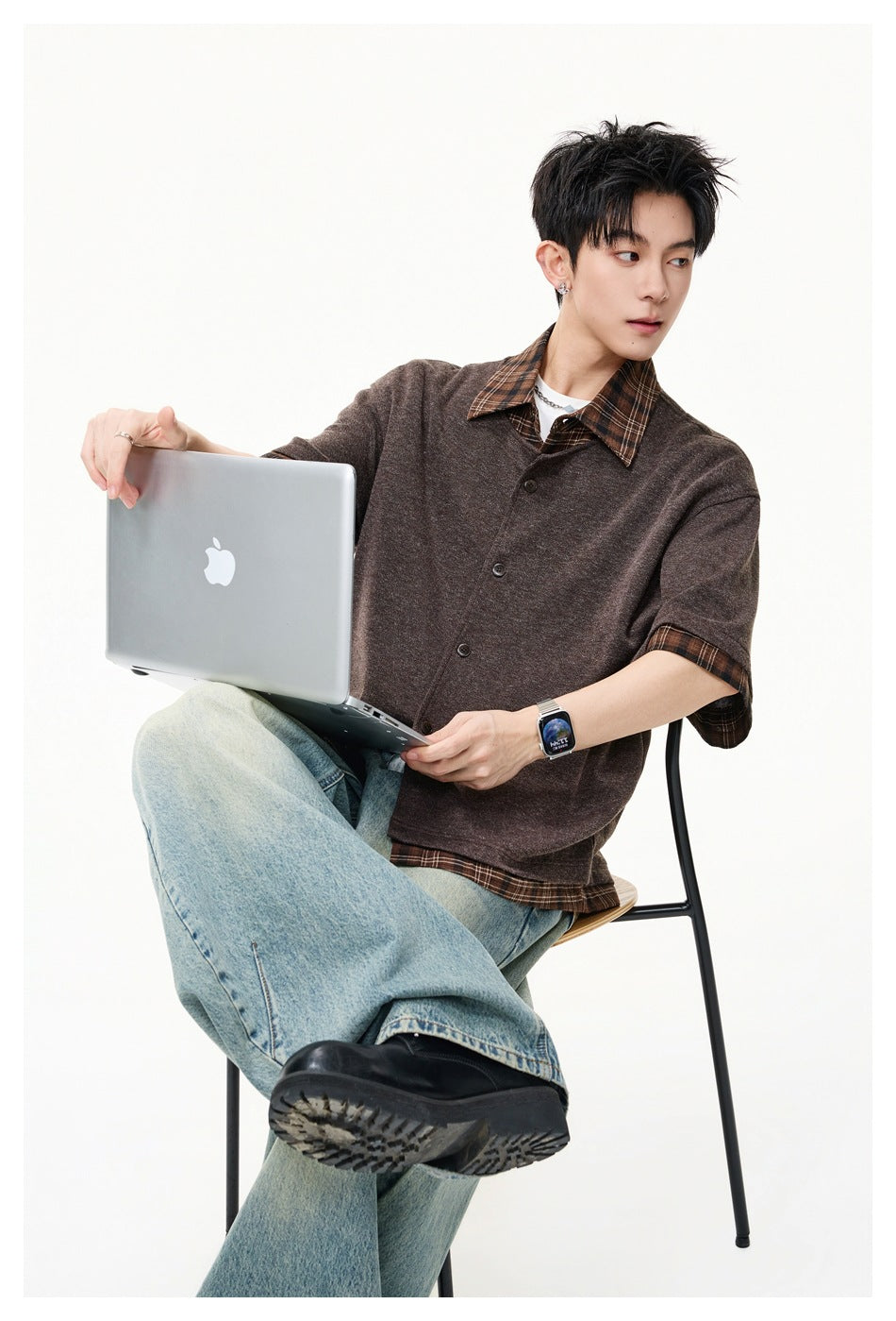 Seated model wearing a brown short-sleeve button-front top with a plaid collar and cuffs over a plaid shirt.